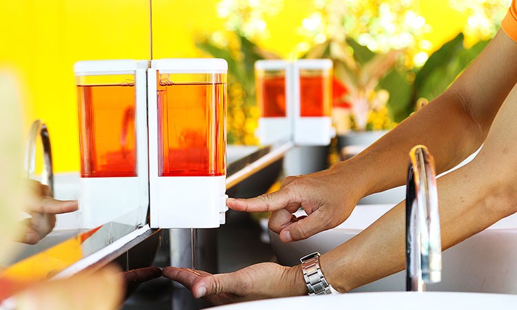 Photo of a pair of hands operating a wall-mounted soap dispenser. PSA tapes can be used to bond these dispensers to the wall. 