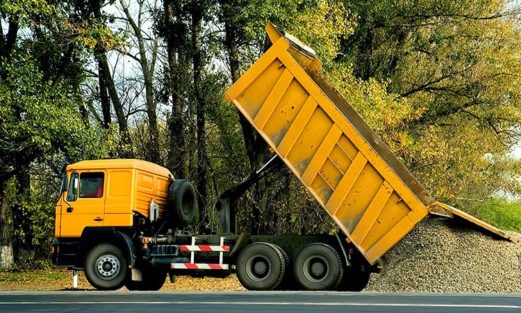 Photo of a large, yellow gravel hauling truck depositing its load of gravel on to the ground by the side of a road.