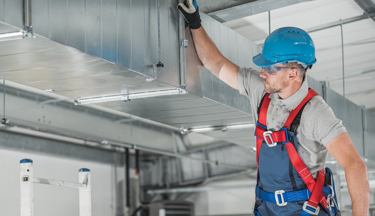 Photo of a technician on a ladder inspecting a section of HVAC ductwork in a commercial building. Our portfolio includes HVAC tapes.